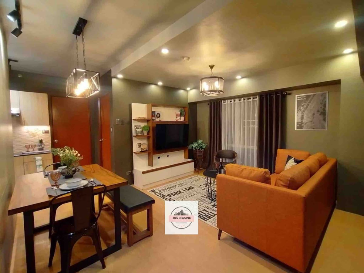 An open living area features a cozy sofa and a modern entertainment unit against a green accent wall. A dining table sits nearby, adorned with place settings. The space is illuminated by warm lighting, contributing to a welcoming atmosphere.