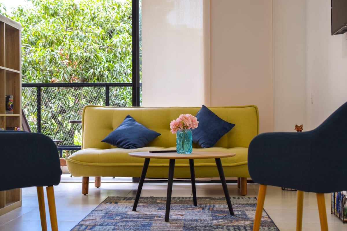 A bright seating area is highlighted by a soft yellow sofa adorned with blue cushions. A floral arrangement in a glass vase sits on a round coffee table, while two dark blue armchairs frame the space. Large windows provide a view of greenery outside.