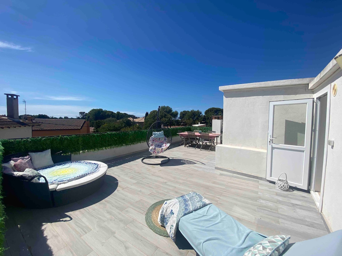 A spacious rooftop terrace features a comfortable lounging chair and a cozy round seating area. Sunlight illuminates the area, surrounded by greenery and offering views of the rooftops. A dining table set for outdoor meals is visible, enhancing the relaxation space.