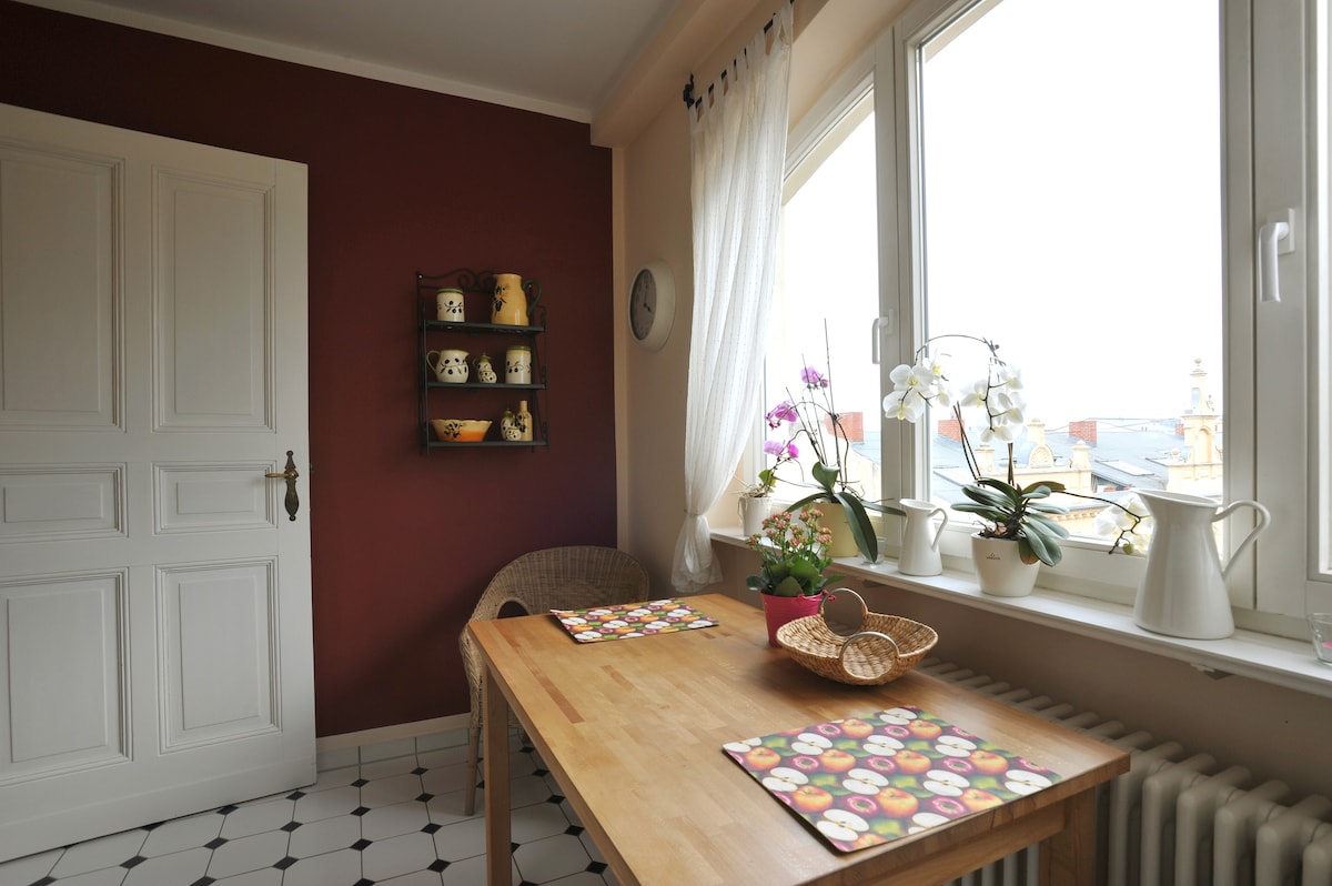 A bright kitchen corner features a wooden table set with patterned placemats. Natural light enters through large windows, illuminating a floral arrangement and potted plants. The wall is adorned with a display shelf holding pottery, while the tiled floor enhances the overall warmth of the space.