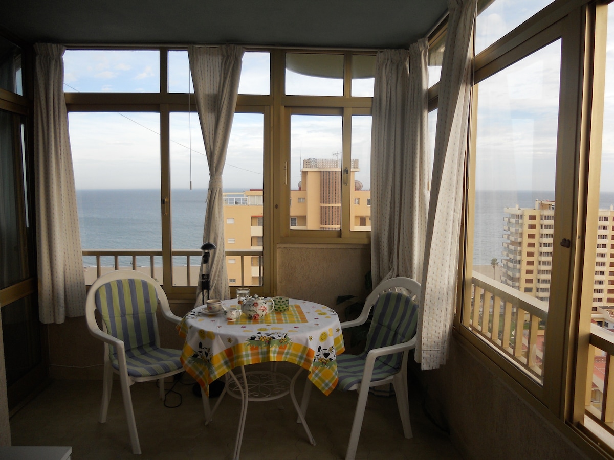 A sunlit glass-enclosed terrace features a circular table set with a checkered tablecloth and two white chairs. The view showcases the sea and nearby buildings through large windows, creating a bright and inviting atmosphere.
