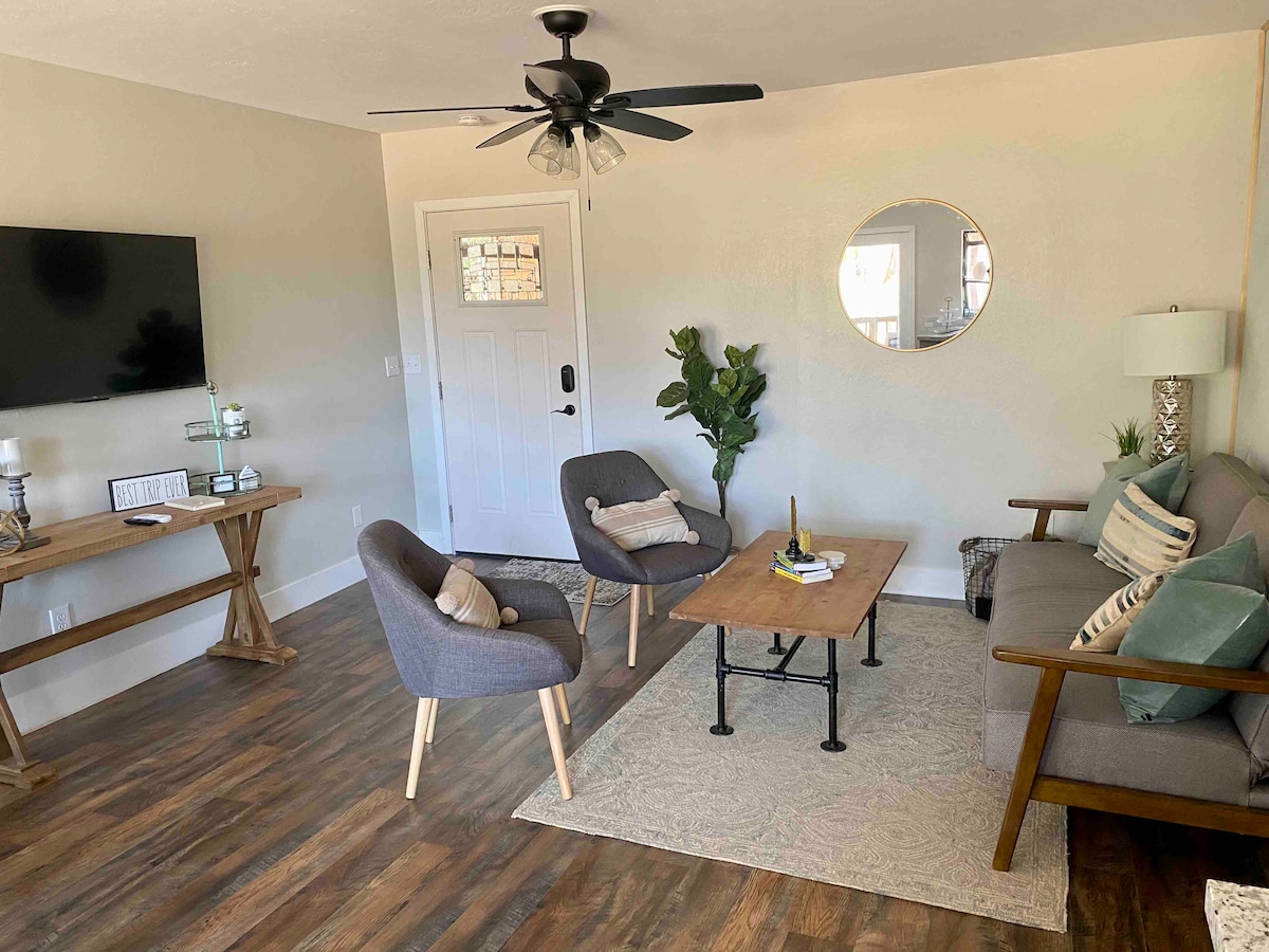 The living area features a cozy layout with a comfortable sofa, two accent chairs, and a coffee table on a textured rug. A wall-mounted TV is visible, along with a large mirror and potted plant, offering a welcoming ambiance.