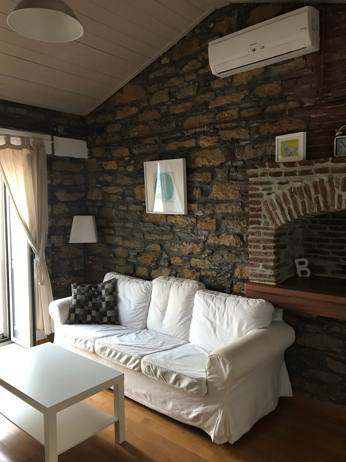 A cozy seating area features a white slipcovered sofa adorned with a patterned cushion. A small white coffee table sits in front of the sofa. Exposed stone walls and a rustic brick fireplace create a textured backdrop. Natural light streams in through the curtains beside a glass door.