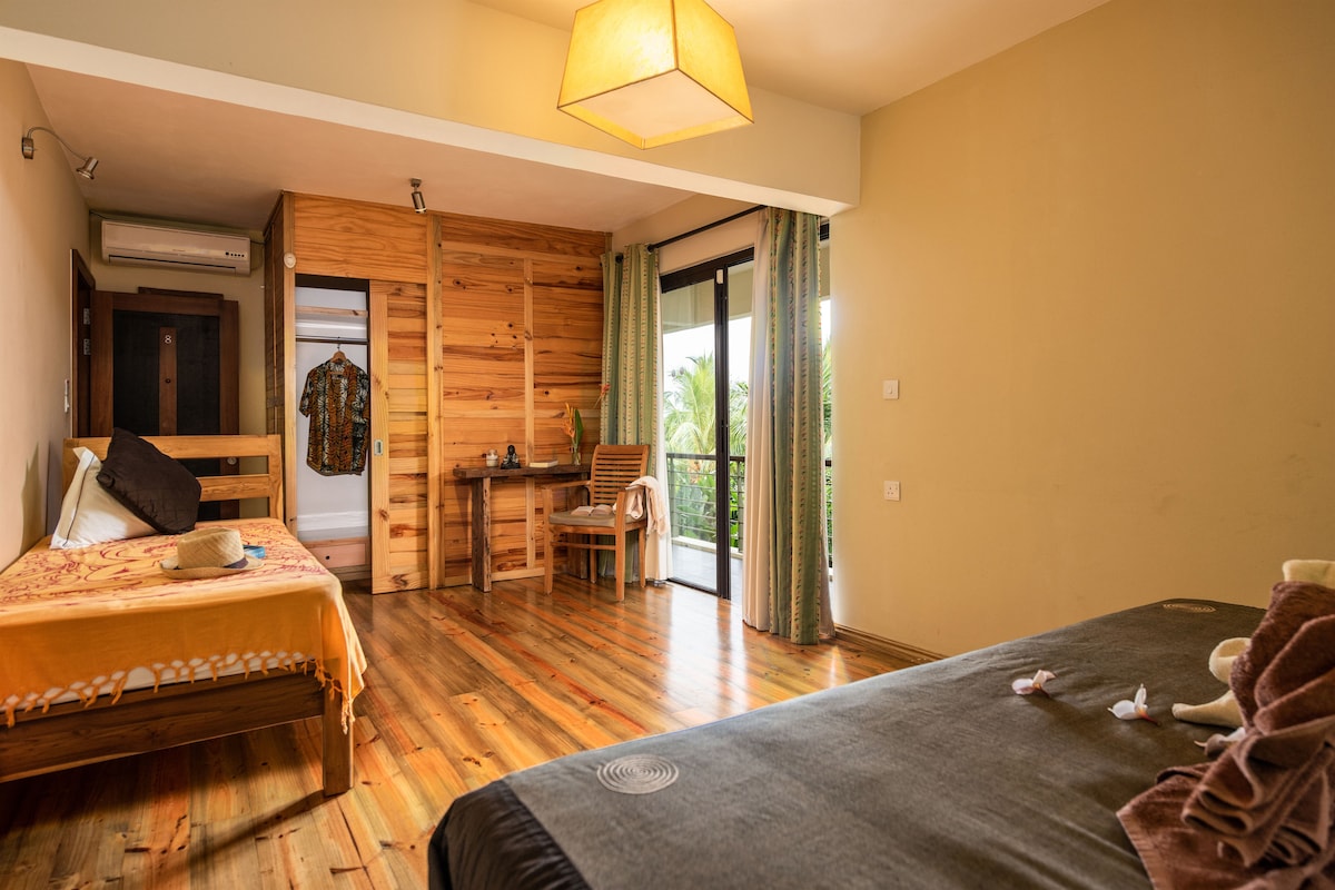 A bright room features wooden flooring and warm-toned walls. A spacious bed is situated beside a light-filled sliding door that opens to a balcony. A wooden desk and chair are positioned against a wall, while decorative accents, such as towels and soft petals, are arranged thoughtfully on the beds.