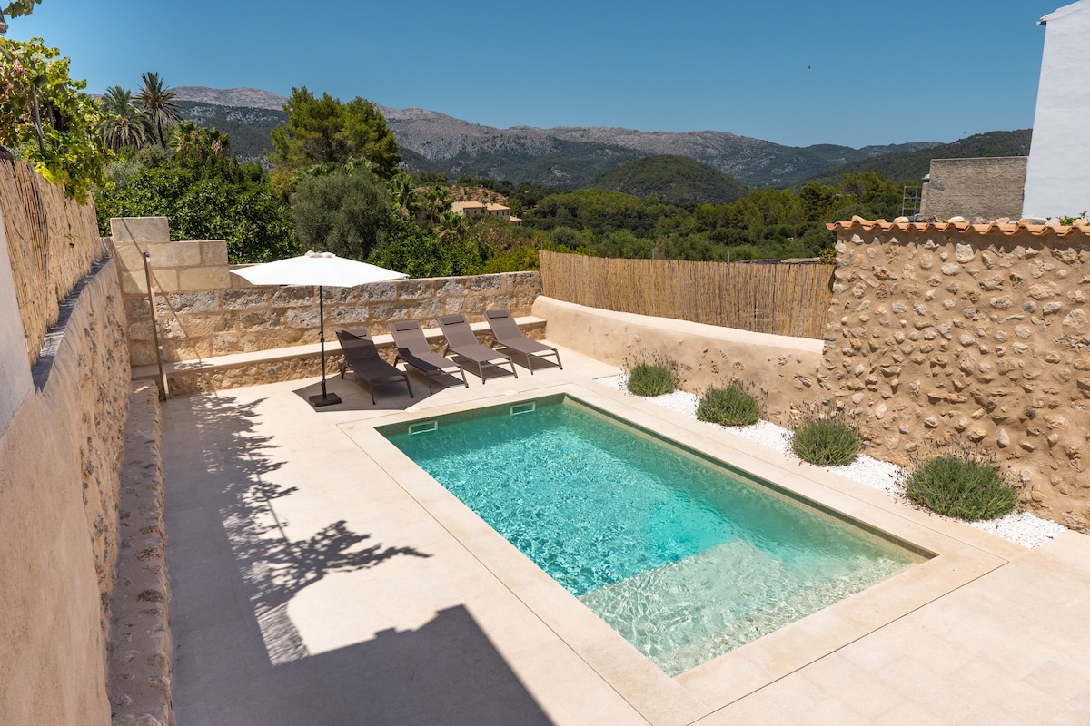 An outdoor area features a rectangular swimming pool surrounded by a spacious sun terrace. Four loungers are positioned under a large umbrella, providing shade. The backdrop showcases gentle hills and greenery, framed by a rustic stone wall that enhances the serene ambiance.