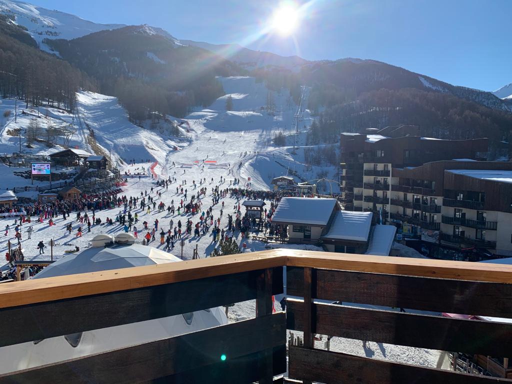 The view from the balcony overlooks a bustling snow-covered ski slope, where skiers and snowboarders can be seen enjoying the day. Mountain ranges rise in the background under a bright sun, casting light over the lively scene below.