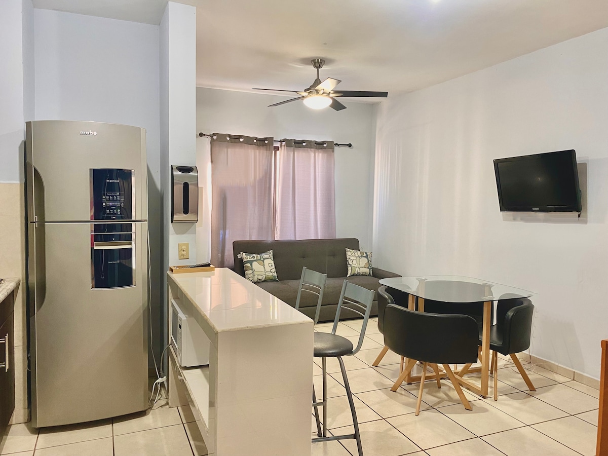 A modern kitchen area features stainless steel appliances, including a refrigerator. A circular dining table with black chairs is positioned near a comfortable sofa. Natural light enters through window coverings, and a ceiling fan is mounted above, enhancing airflow in the space.