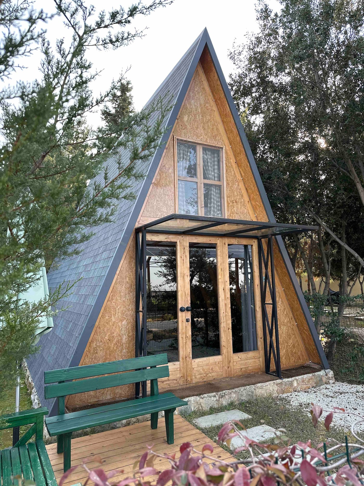 An A-frame structure showcases a combination of wood and glass elements. The entrance features double doors beneath a black overhang, framed by greenery and a wooden deck. A green bench is positioned in front, enhancing the outdoor space.