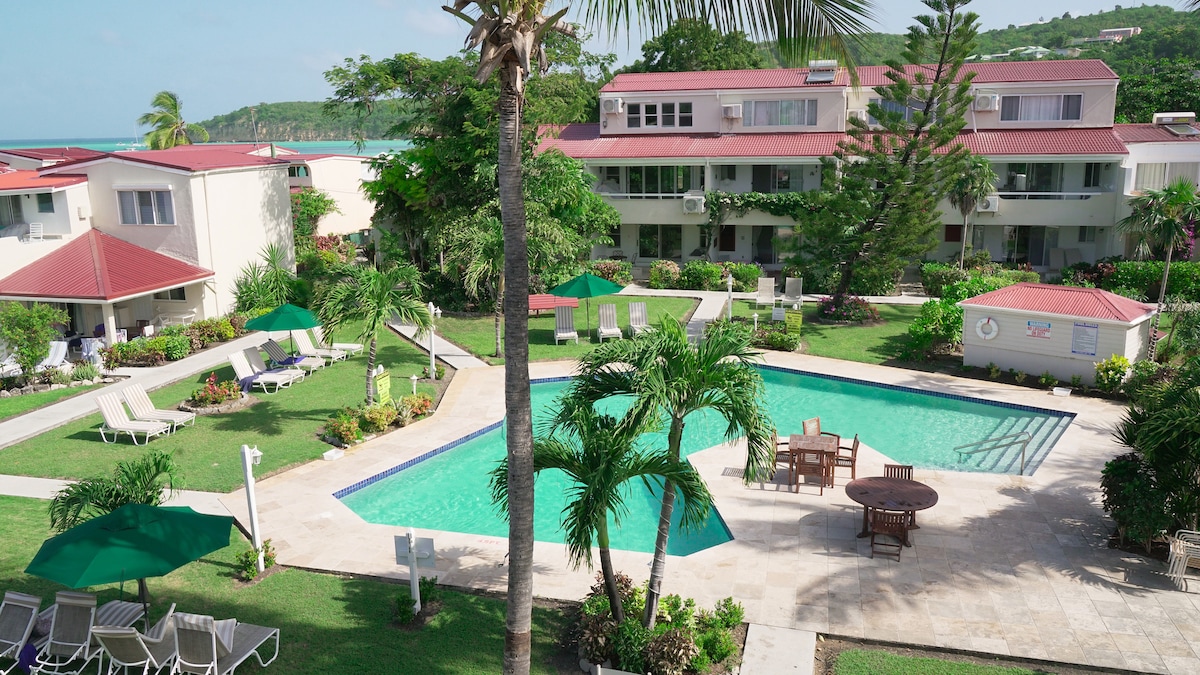 A lush tropical garden surrounds a large swimming pool, featuring sunbeds and dining furniture. The pool area is positioned between two multi-story buildings, with vibrant greenery and palm trees adding to the tranquil atmosphere. The distant view hints at nearby beaches.