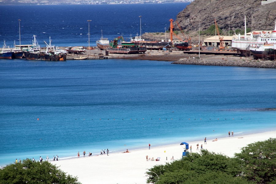 A tranquil beach scene features soft white sand and clear blue waters. Small groups of people are seen walking along the shore, while a beach umbrella provides shade. In the background, fishing boats are anchored near a rocky coastline, framed by a gentle hillside.