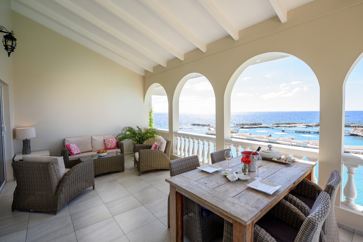 A spacious terrace offers a dining area with a wooden table set for meals, surrounded by comfortable seating. Arched openings frame views of the Caribbean Sea and outdoor area. The setting is complemented by soft cushions in neutral tones and potted greenery.
