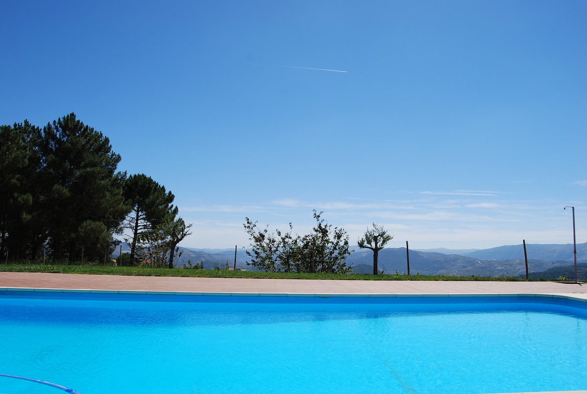 A clear blue swimming pool reflects the sky, framed by a spacious deck and surrounded by lush trees. Scenic hills and distant valleys create a tranquil backdrop, enhancing the serene outdoor environment.