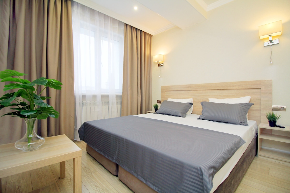 A bright bedroom features a spacious bed paired with neutral-toned bedding. Soft light from a window filters through sheer curtains, enhancing the serene environment. A small bedside table with a plant adds a touch of greenery, while wall sconces provide additional illumination.
