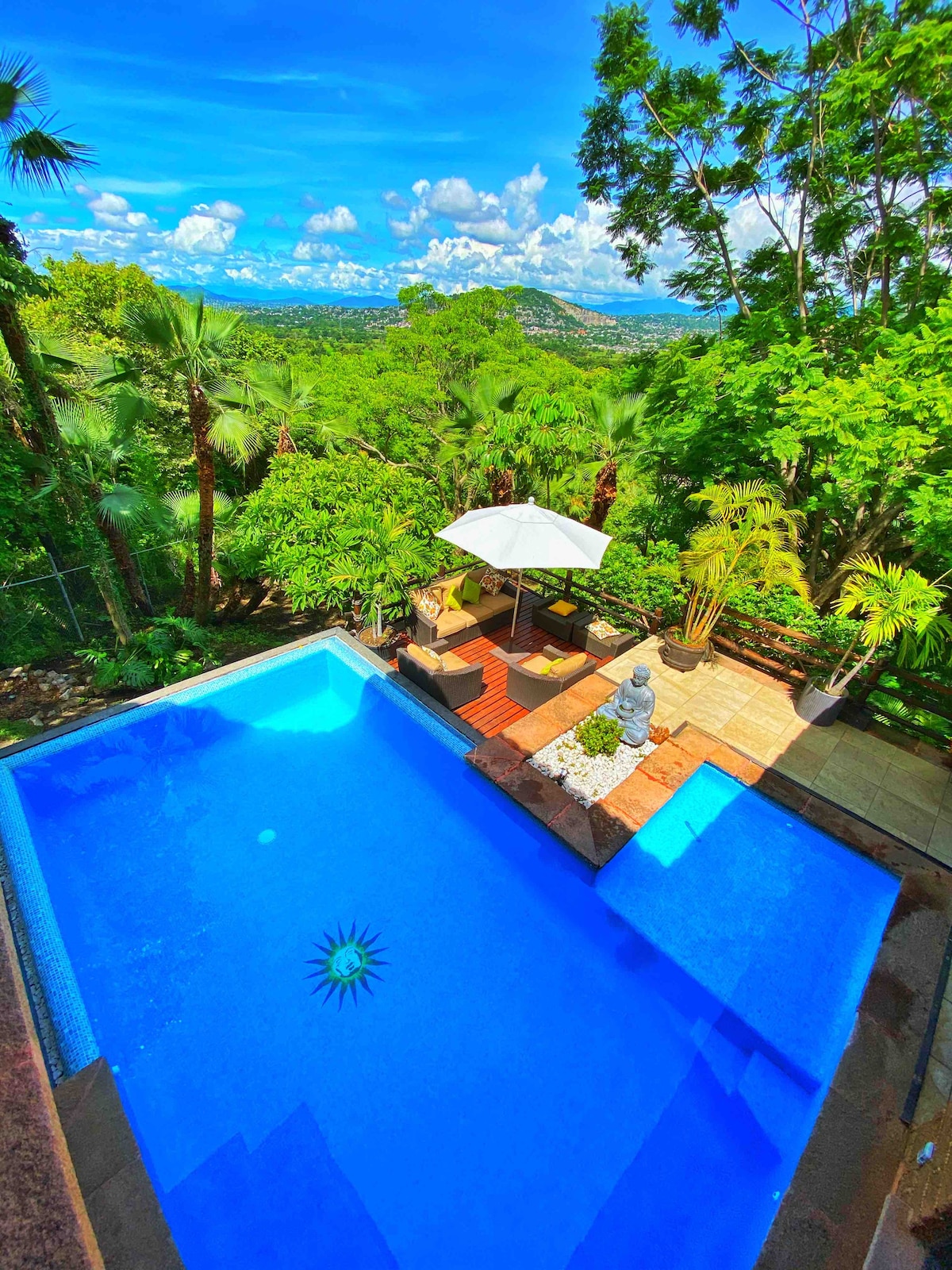 A private heated pool is surrounded by lush greenery, with a patio area featuring a sun umbrella and comfortable seating. The landscape offers views of distant hills and a vibrant sky, creating a serene outdoor oasis.