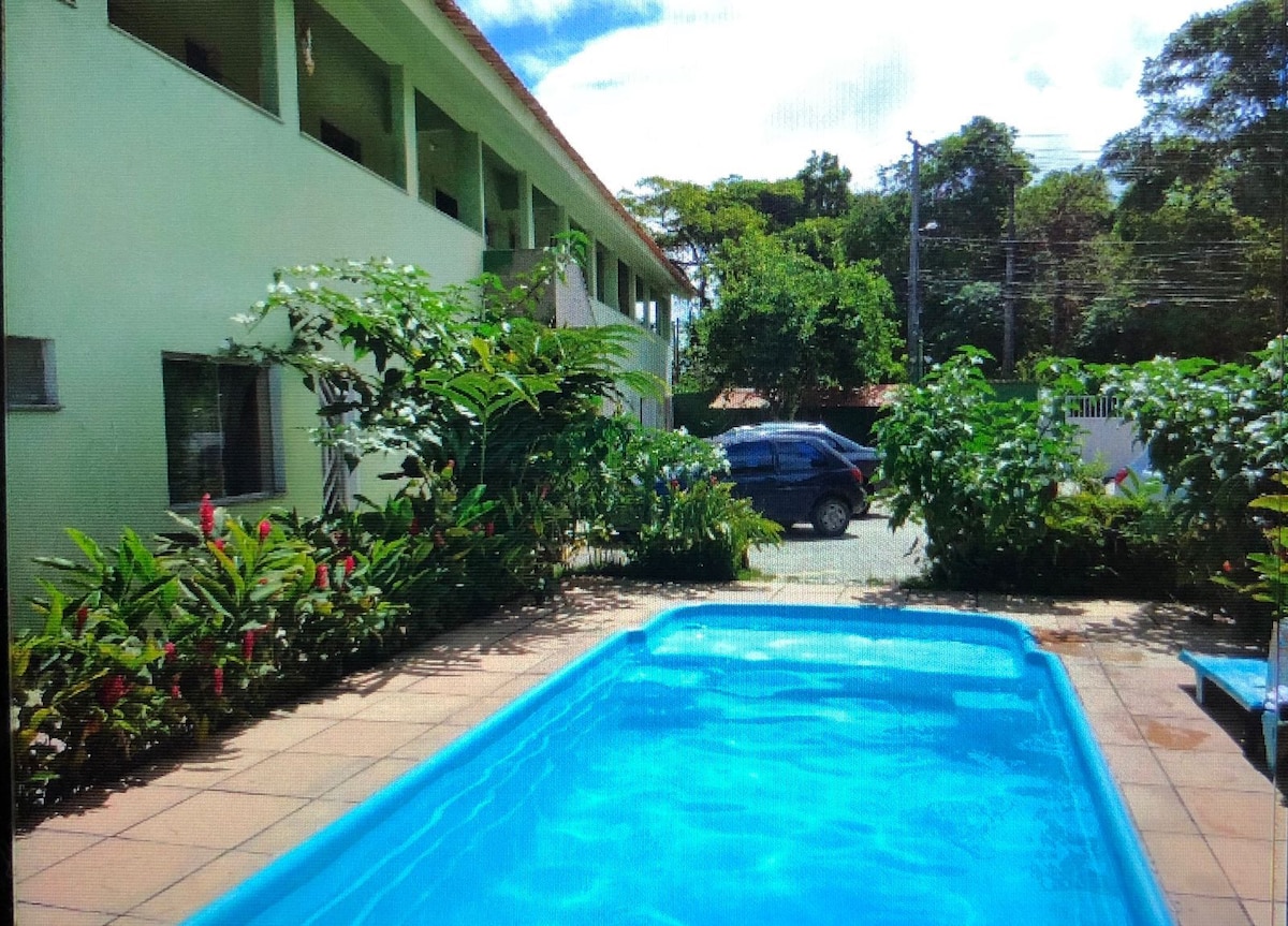 A clear blue pool is surrounded by lush greenery and vibrant plants. A paved area lies beside the pool, featuring a small seating area. The building in the background is two stories tall, with large windows and is set within a natural environment.