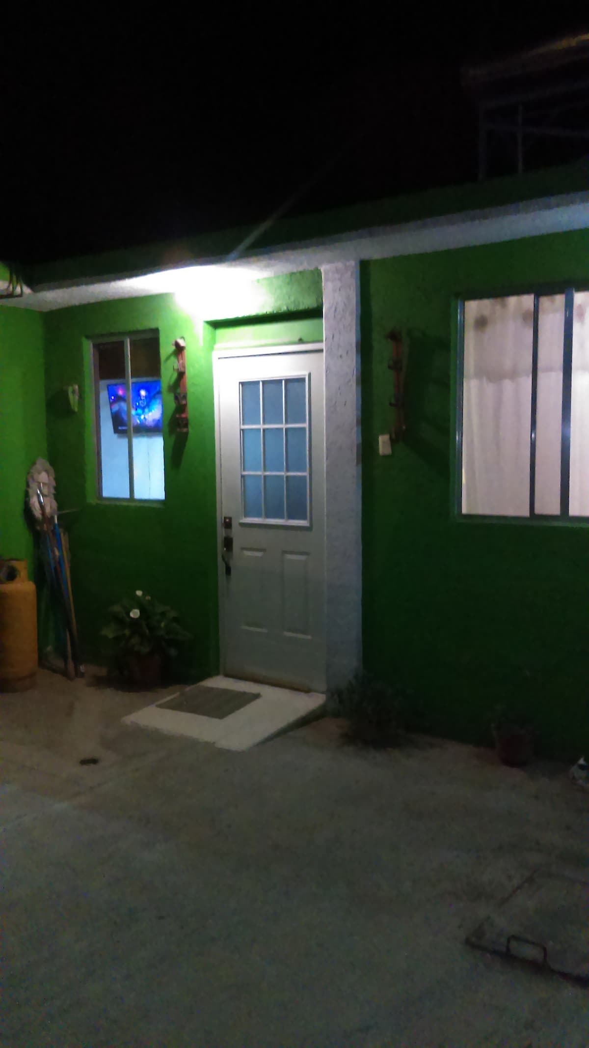 The image shows a vibrant green exterior of a house, featuring a white door with glass panels. A large window is visible beside the door, surrounded by decorative elements. Soft lighting illuminates the entrance, creating a welcoming atmosphere at night.