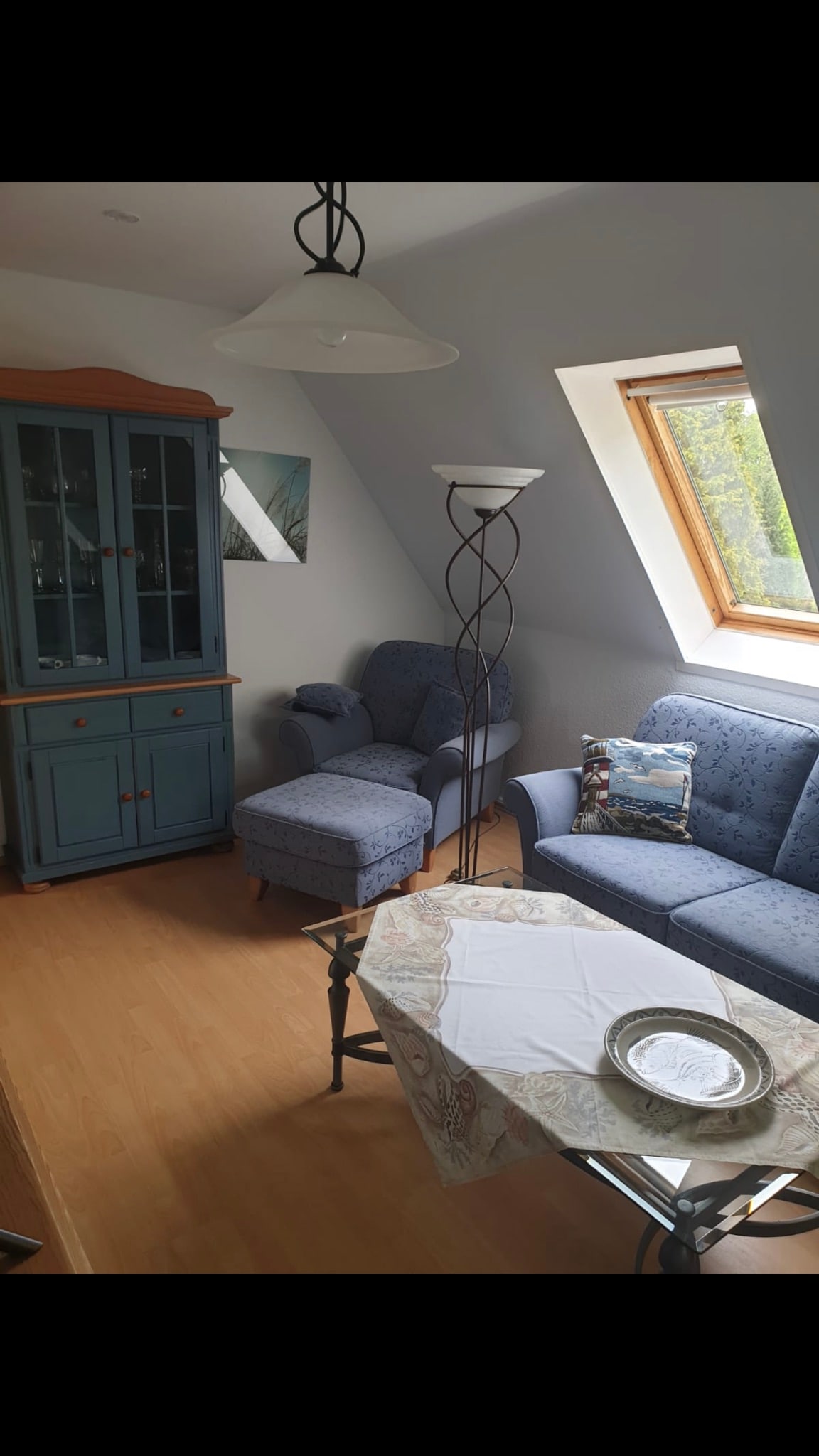 A comfortable living area features soft blue armchairs and a matching couch, accompanied by a glass-top table. Natural light enters through a skylight above, illuminating wooden floors and a sideboard displaying various decorative items. A decorative pillow adds a touch of color to the seating.