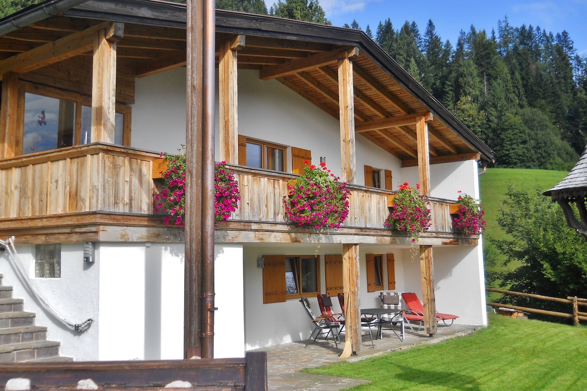 The chalet features a spacious balcony adorned with vibrant flower boxes. Sun loungers are positioned on the stone patio, while lush greenery and forested hills provide a serene backdrop. Large windows allow natural light to fill the interior spaces.