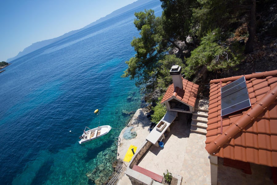A serene coastal view is captured, showcasing a boat moored nearby and clear blue waters. A stone terrace with a red-tiled roof is visible, complete with a barbecue area. Lush green pine trees frame the scene, enhancing the natural surroundings.