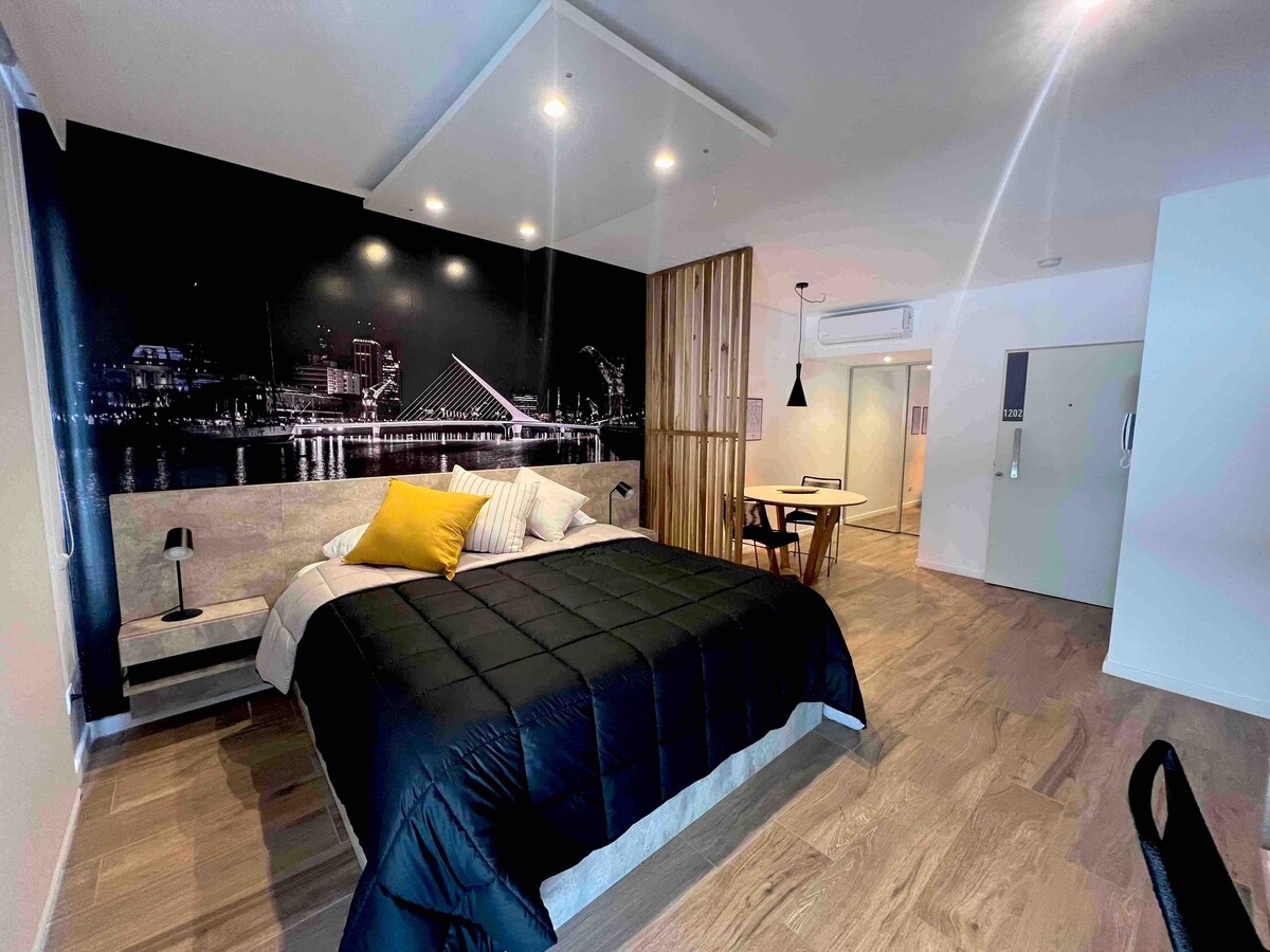 A spacious sleeping area features a large bed with a dark quilt and decorative pillows. A striking cityscape mural serves as a backdrop, while natural light enters from a nearby window. A dining table can be seen in the distance, completing the modern aesthetic.