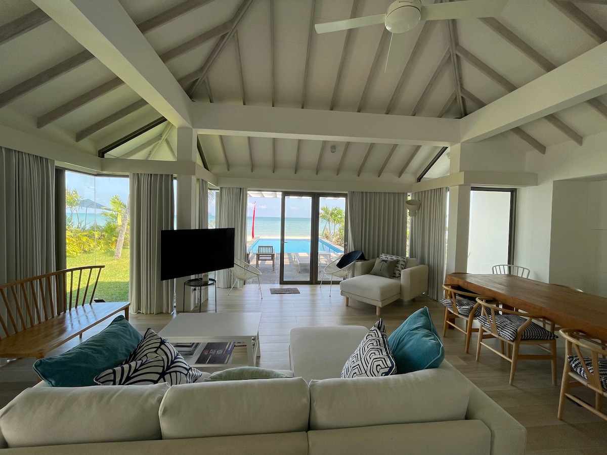 A spacious living area features large glass windows that provide views of the beach and garden. Light-colored furniture is arranged around a wooden dining table, with comfortable seating options available. High ceilings with exposed beams enhance the airy atmosphere of the space.