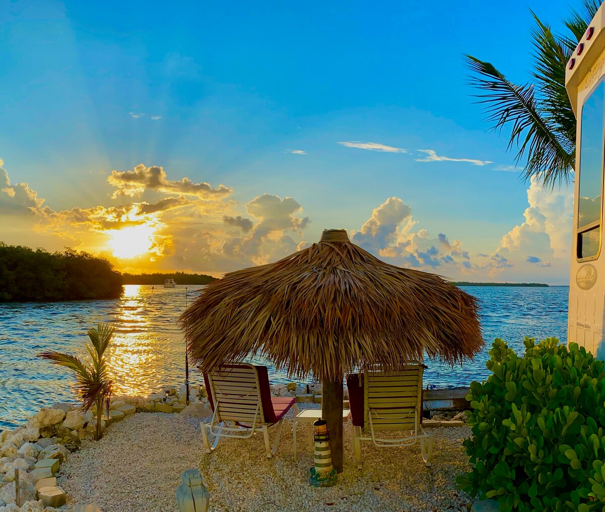 A tiki hut with thatched roofing provides shade over two lounge chairs facing the water. The scene is complemented by vibrant sunlight filtering through the clouds, casting warm tones across the tranquil waterfront and enhancing the natural beauty of the environment.