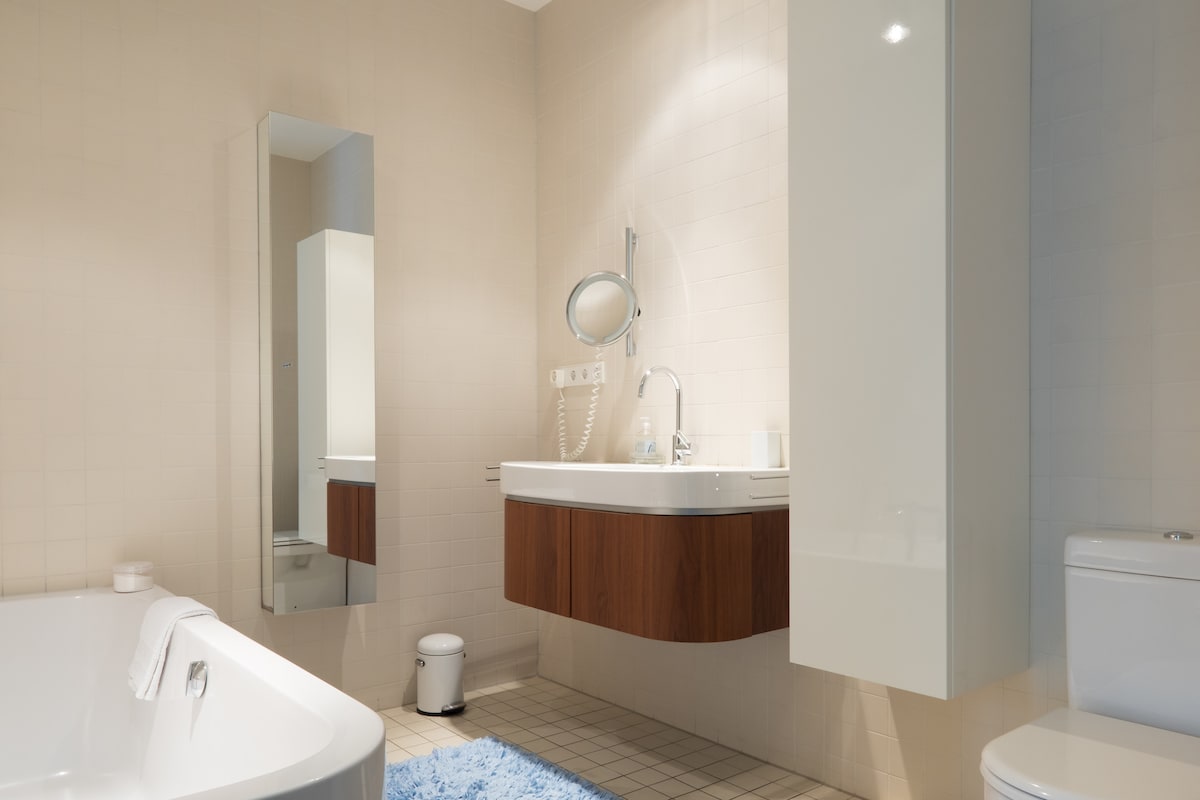 The bathroom features a spacious bathtub with a sleek wooden vanity above it. A large mirror reflects the contemporary design, and a round magnifying mirror is mounted on the wall. Soft blue bath mat complements the neutral tiled floor, enhancing the room's overall ambiance.