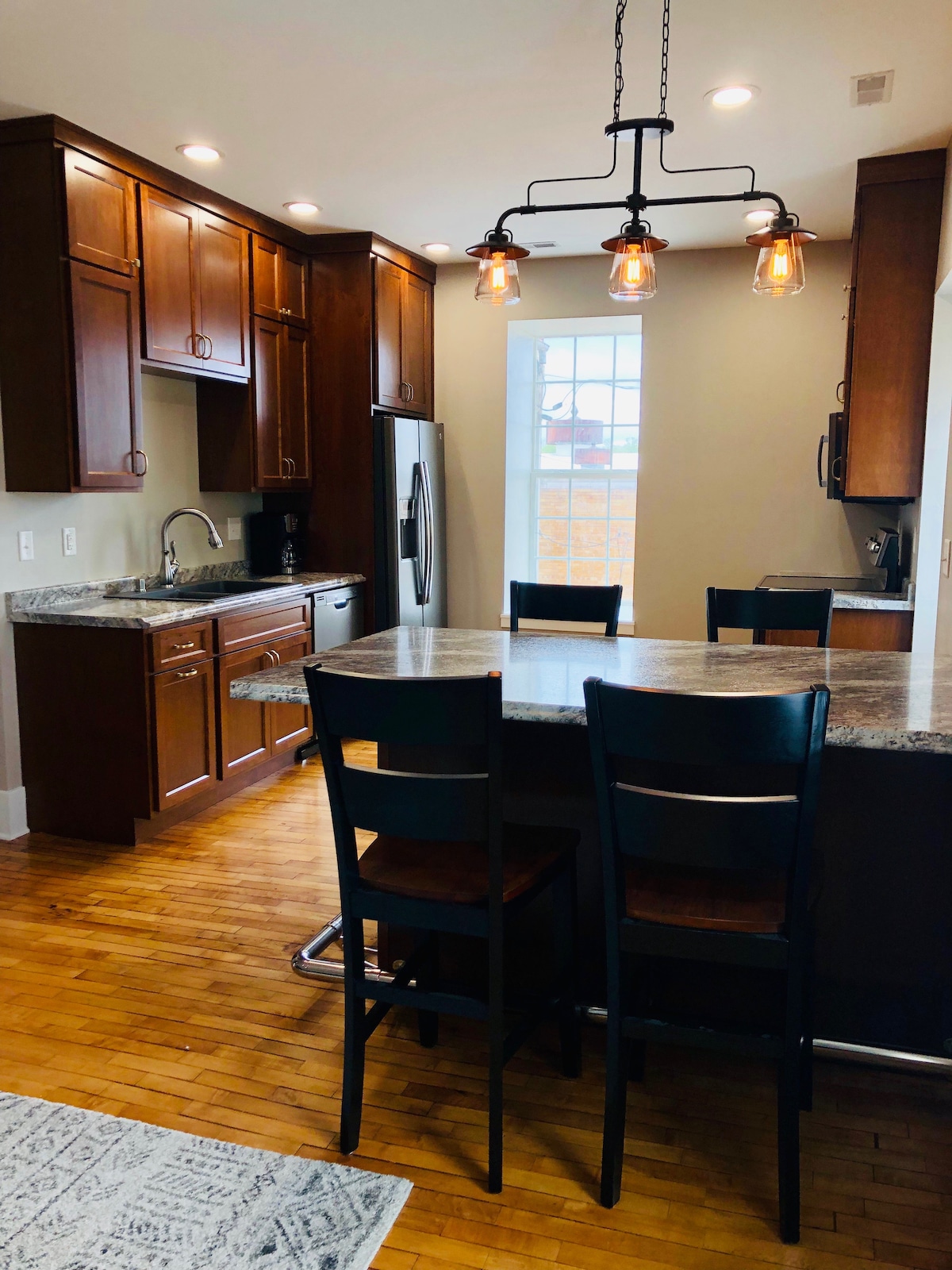 A modern kitchen features dark wood cabinets, stainless steel appliances, and a spacious granite countertop with seating for three. Natural light fills the room through a window, illuminating the warm wooden flooring.