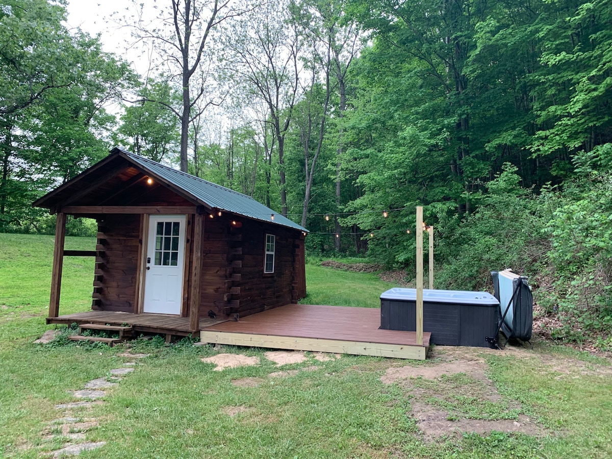 A charming cabin features a wooden exterior and a welcoming white door. A spacious deck is attached, with a hot tub positioned beside it. Surrounding greenery includes trees, creating a peaceful setting. Soft lighting illuminates the area, enhancing the outdoor space.