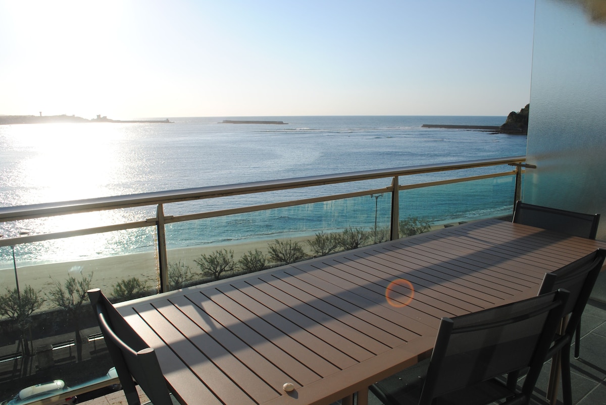 A spacious terrace offers a stunning view of the bay, with a large dining table and several chairs positioned for outdoor dining. The water shimmers in the sunlight, highlighting the peaceful coastal atmosphere.