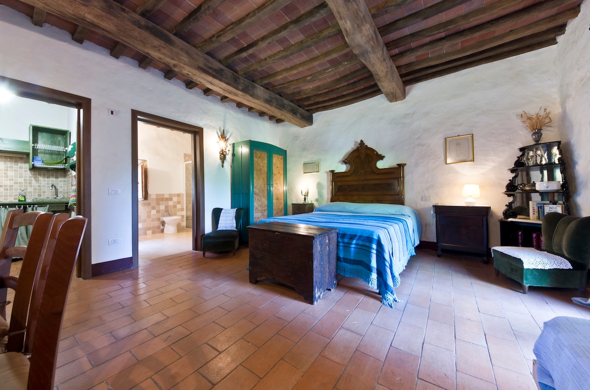 A spacious bedroom features a wooden beam ceiling and terracotta flooring. A large bed with a blue striped blanket is centered in the room. A vintage wooden cabinet and a small green chair are positioned nearby, with a soft light emanating from a lamp on a dark wooden nightstand.