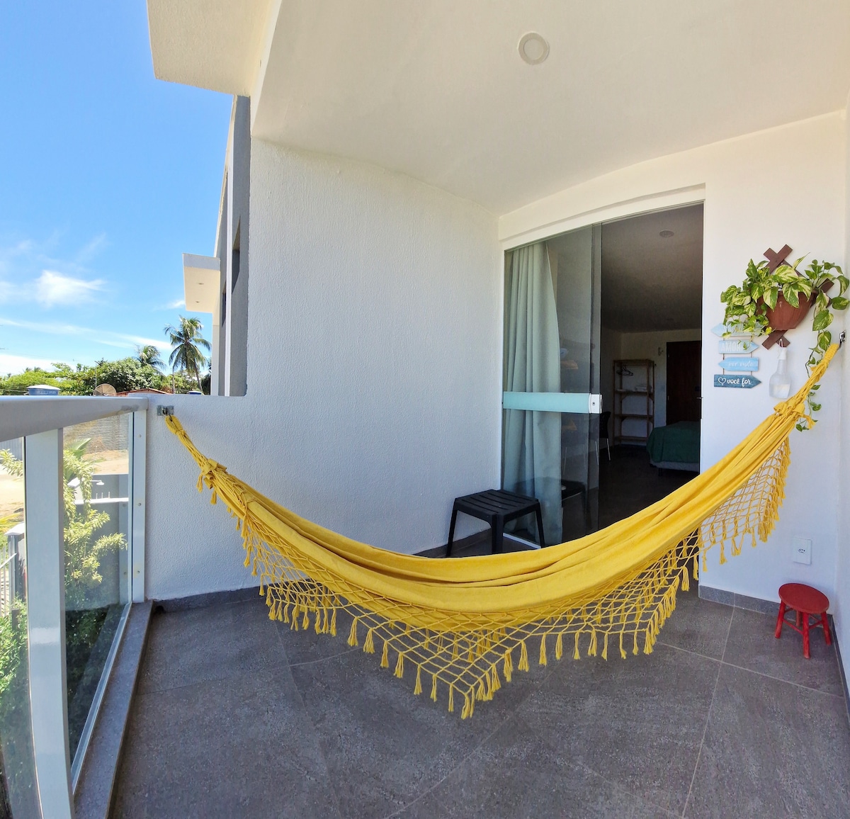 A sunlit balcony features a vibrant yellow hammock gently draped and swaying. A small black table sits adjacent to the hammock, providing a perfect spot for relaxation. Sliding glass doors offer a view into the inviting interior space.