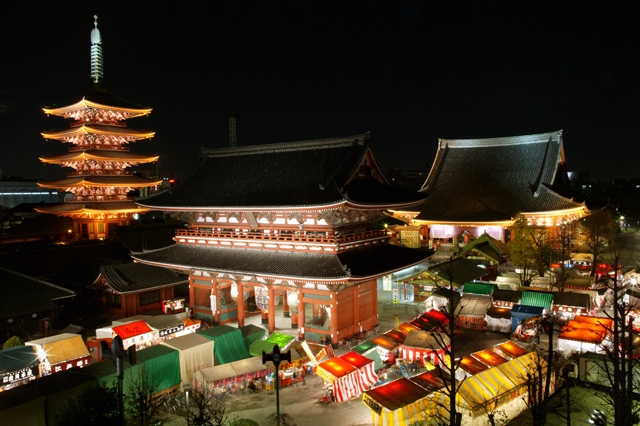 The nighttime view captures the illuminated buildings of a Japanese temple complex. A pagoda is visible alongside traditional structures, with stalls displaying vibrant canopies surrounding them. Soft lights enhance the scene, creating a peaceful atmosphere amid the bustling market below.