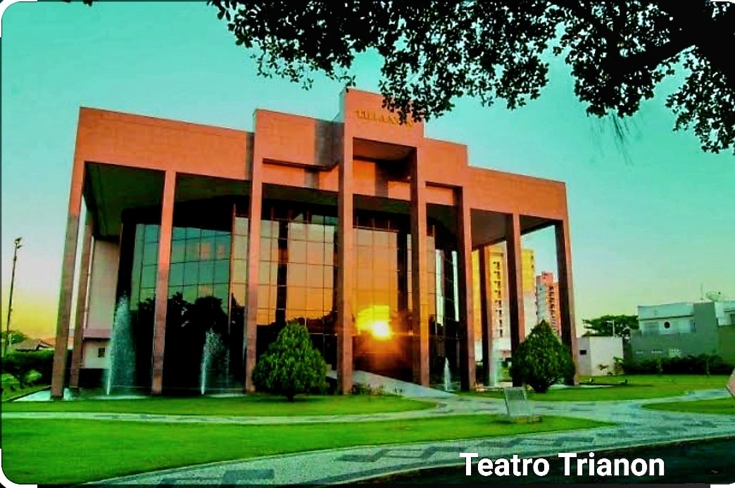 The grand exterior of Teatro Trianon is highlighted, featuring large glass windows that reflect the sunset. Surrounding greenery and manicured lawns enhance the open space, while the architectural design offers a modern aesthetic with geometric shapes.
