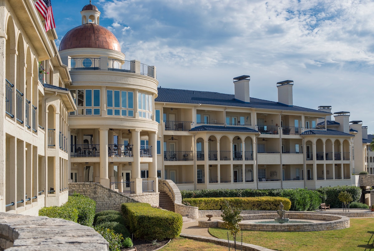 The image features a resort-style structure with a distinctive dome atop the central building. The architecture highlights an elegant blend of modern and classic design, with multiple balconies and expansive windows. A lush, landscaped area is visible in the foreground, enhancing the serene surroundings.