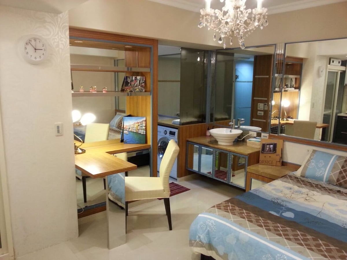 A well-lit living space includes a cozy bed with patterned linens, a wooden desk and chair, and a modern chandelier. A small dining area is present, along with a kitchen space featuring a washer. Mirrors enhance the brightness of the room.