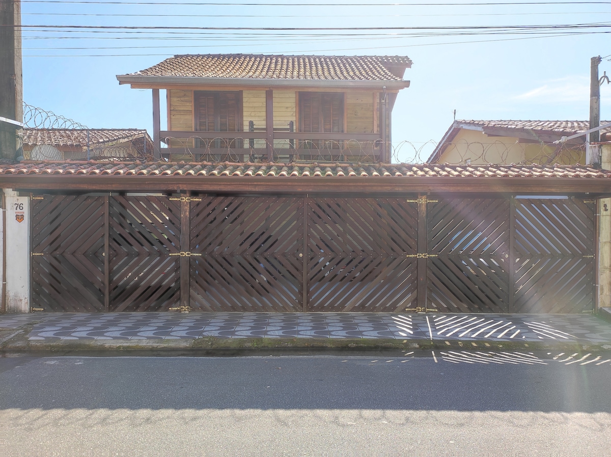 The front exterior of the property features a wooden gate with a geometric design, leading to a spacious driveway. Above, a second-story house with a covered balcony is visible, surrounded by a tiled pathway and residential structures.