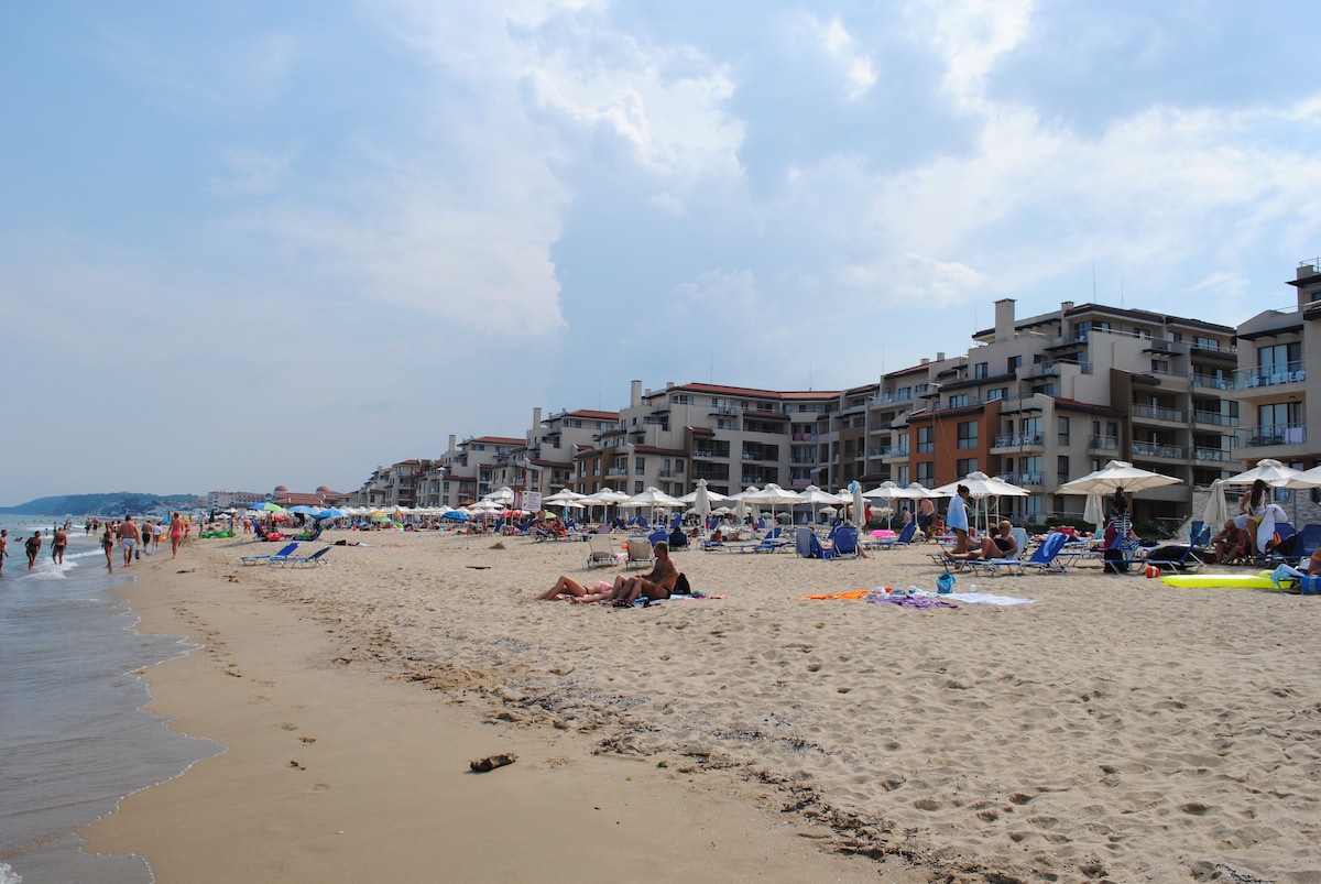A sandy beach scene is depicted, featuring clusters of sunbathers on colorful towels and lounge chairs. Sun umbrellas provide shade along the shoreline. In the background, multiple resort buildings are visible, complemented by a varied sky highlighted by soft clouds.