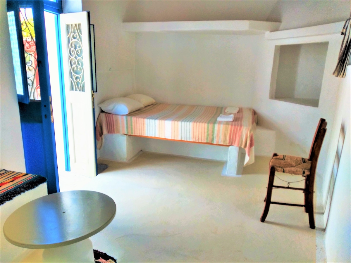 A bright room is shown featuring a single bed with a striped blanket, set against white walls. A wooden chair is positioned beside a small round table. The entrance features blue doors, allowing natural light to flow into the space.