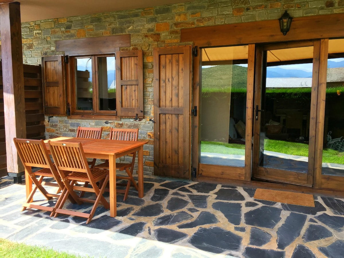 An outdoor seating area features a wooden table surrounded by four folding chairs. The space is framed by stone walls and wooden shutters, providing a rustic charm. Glass doors lead into the living area, and lush greenery is visible beyond the patio.