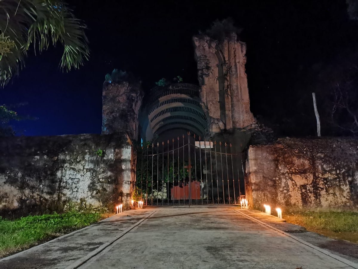 An arched entrance is framed by tall stone pillars, illuminated by flickering lights along the pathway. The dark silhouette of a historical structure rises in the background, surrounded by lush greenery under a night sky.