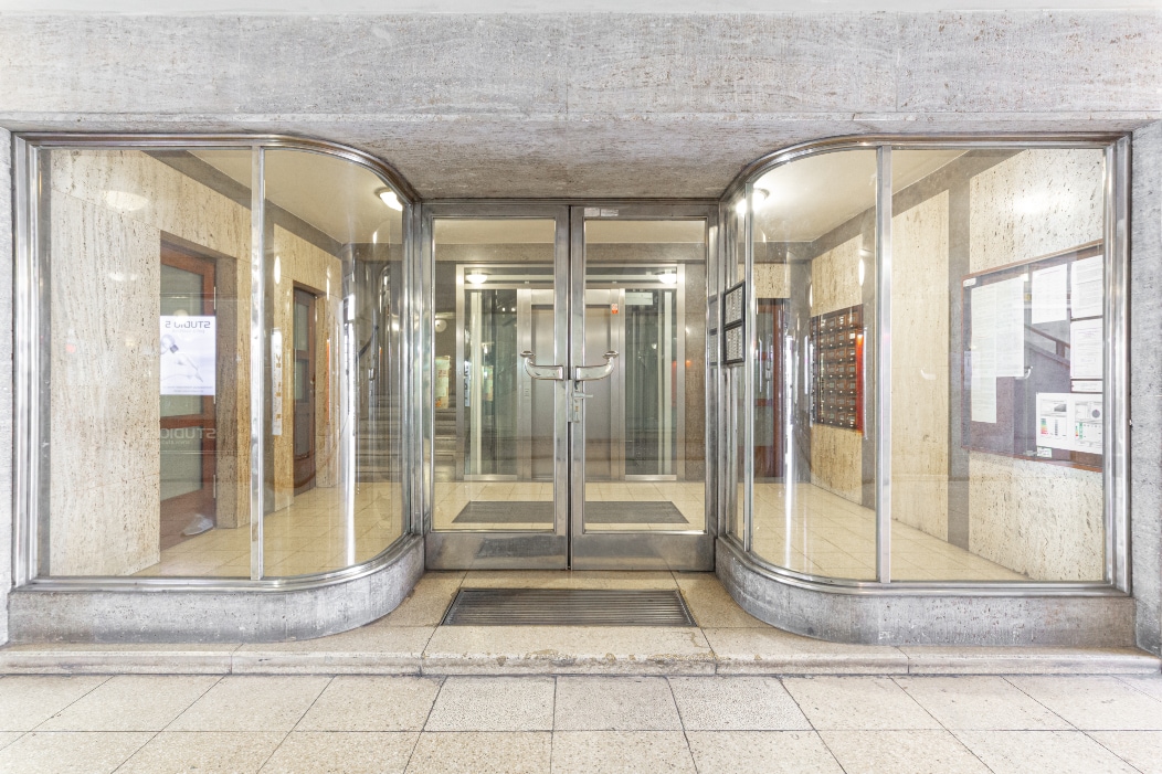 The entrance to the Victoria Palace building features a modern glass façade with curved doors. Inside, a spacious lobby can be seen, highlighted by marble walls and polished flooring. Elevators are accessible, alongside mailboxes arranged on the side walls.