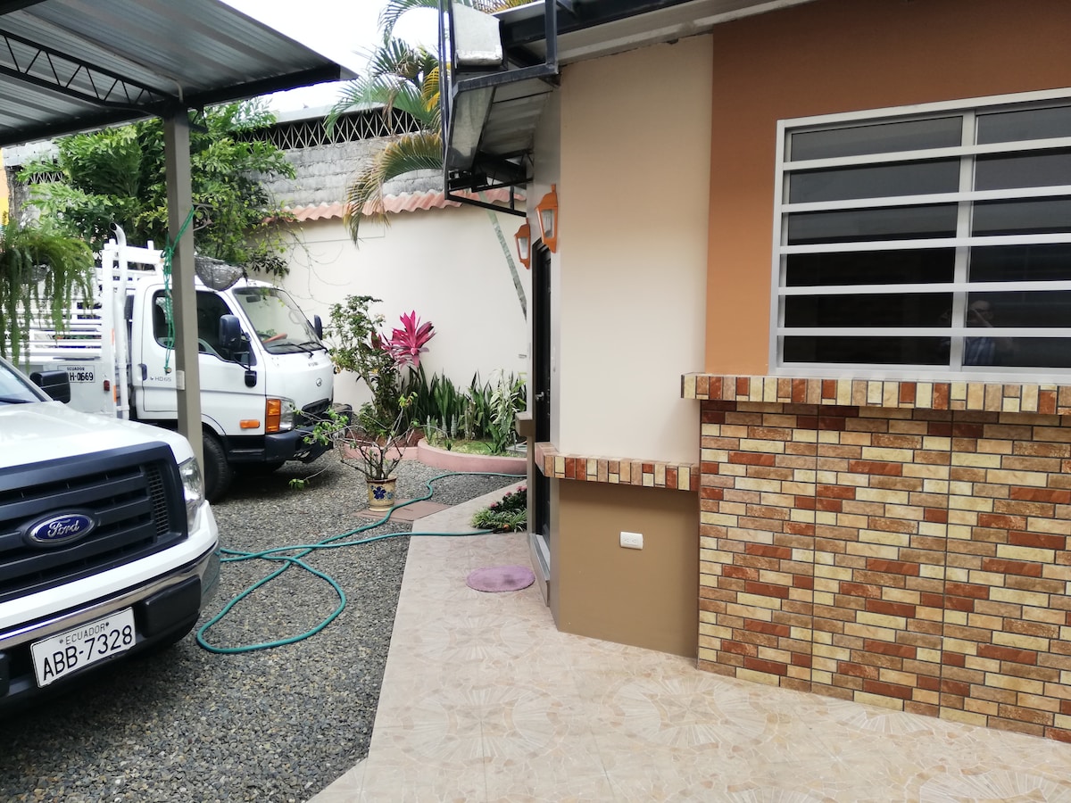 An outdoor area is showcased, featuring a combination of tiled surfaces and decorative stonework. Lush plants and flowers add greenery, while a vehicle is parked nearby, emphasizing accessibility. A water hose is visible beside the building, hinting at maintenance of the surrounding landscape.