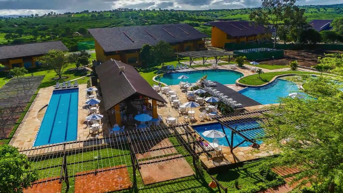 An aerial view reveals a well-maintained outdoor pool area featuring two swimming pools, sun loungers, and umbrellas. Surrounding greenery enhances the setting, with pathways leading to a bar and shaded seating areas. The modern buildings are positioned in the background, providing a scenic backdrop.