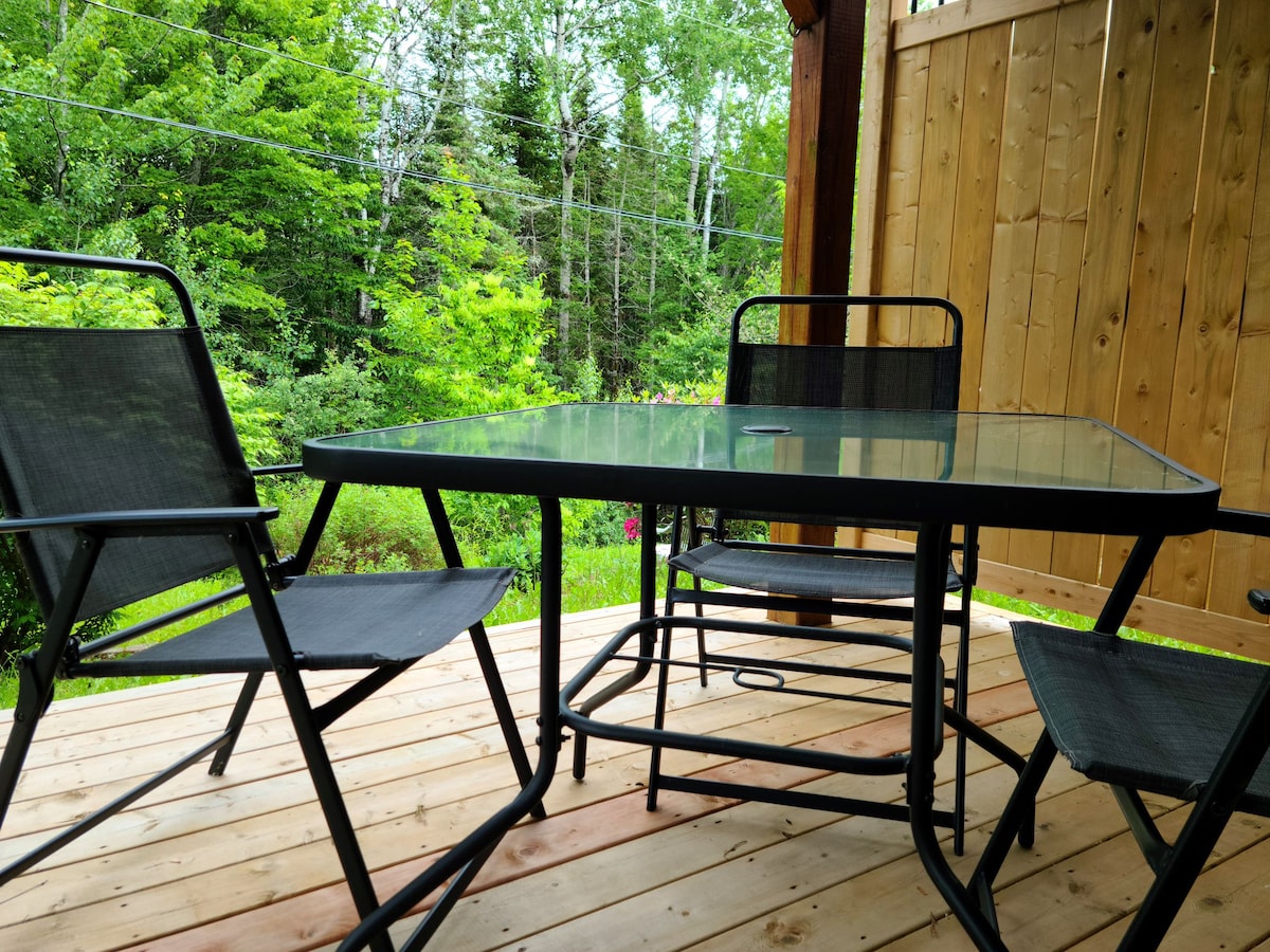 A wooden deck space is featured, equipped with a glass-top table surrounded by four black metal chairs. Lush greenery and trees are visible in the background, creating a tranquil outdoor setting.