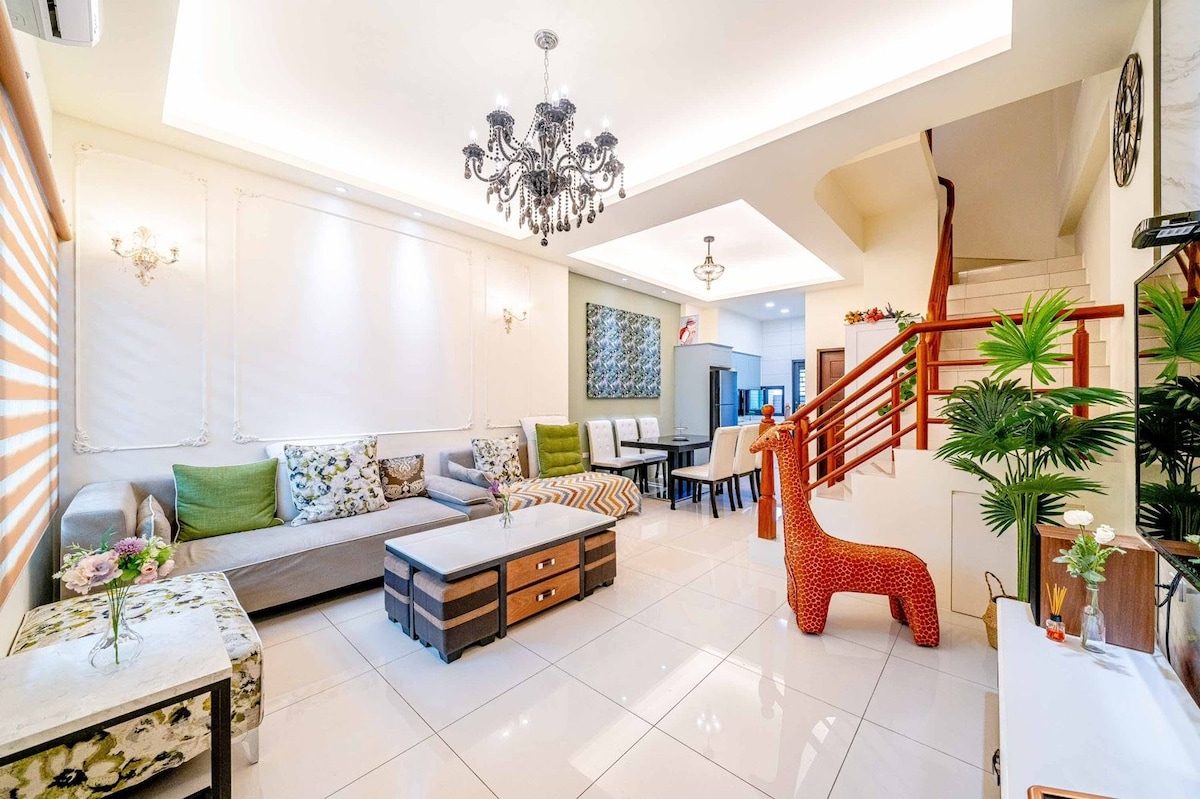 A stylish living area is highlighted by a decorative chandelier and a wooden coffee table. A combination of patterned and solid-colored cushions adorns the spacious sofa. A staircase leads to the upper level, while a dining area is visible in the background, framed by soft lighting.
