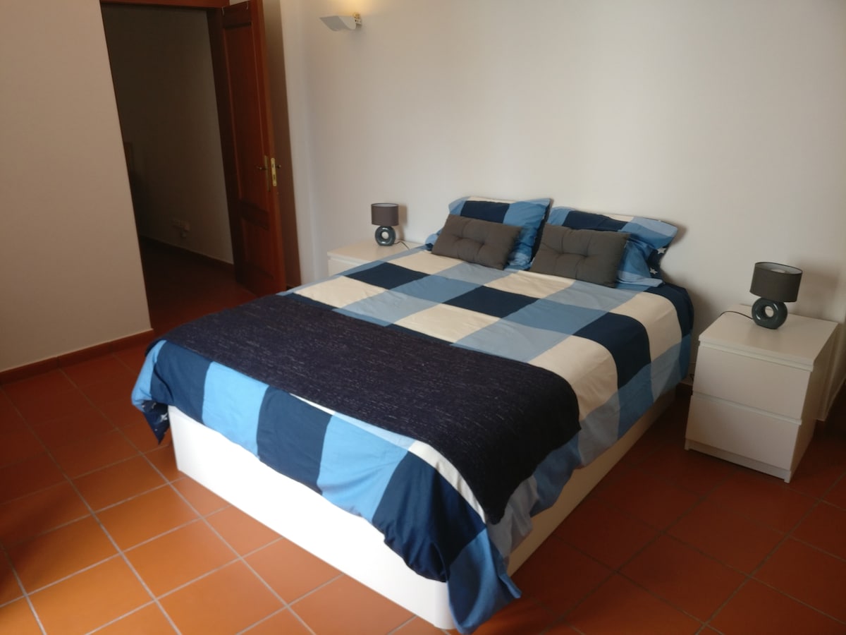 A spacious bedroom features a large bed draped with a blue and white patterned coverlet, complemented by two small nightstands with bedside lamps. Warm sunlight illuminates the tile floor, enhancing the room's inviting atmosphere.