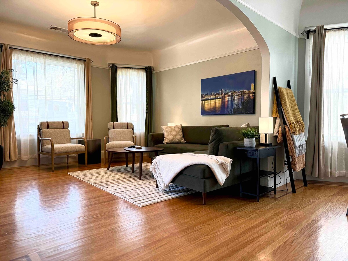 The living room features a modern sofa and two comfortable armchairs arranged invitingly on a hardwood floor. Large windows allow natural light to illuminate the space, accented by soft drapes. A coffee table sits at the center, with a decorative throw laid over the arm of the sofa.