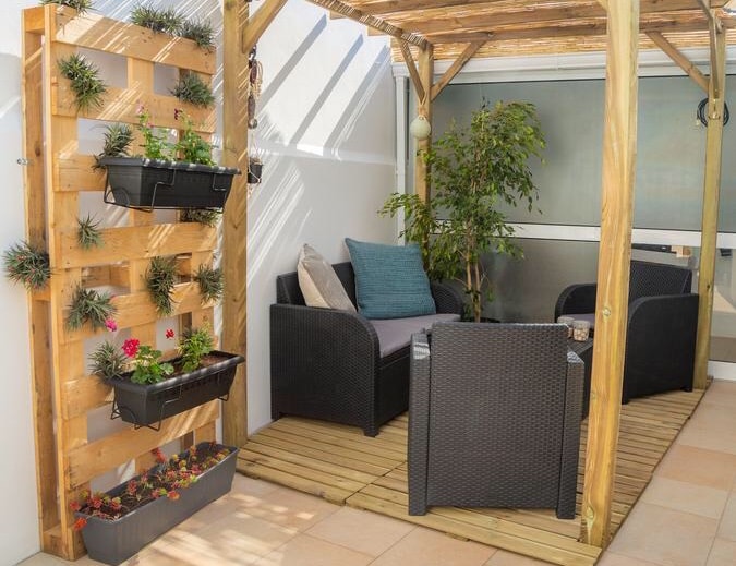 A covered terrace features a wooden framework with seating arrangements, including two armchairs and a small couch adorned with a cushion. Vertical planters filled with various plants and flowers are mounted on the wall, providing a serene environment for relaxation.