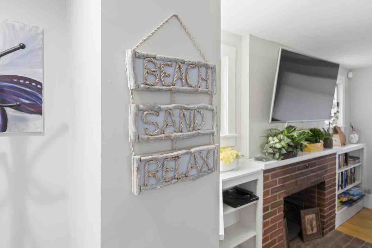 A decorative sign hangs on the wall, displaying the words 'Beach', 'Sand', and 'Relax' in a coastal style. A television mounted above a brick fireplace is visible, flanked by greenery and neatly arranged items on shelving, creating a comfortable living area.